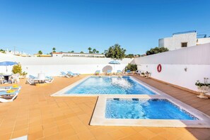 Ein rechteckiger Pool im Freien mit Fliesen umrandet, umgeben von Liegestühlen und Sonnenschirmen, sowie eine weiße Wand mit einem Rettungsring.