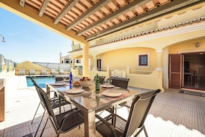 A covered outdoor dining area with a pool, dining tables, chairs, and a view of the pool.