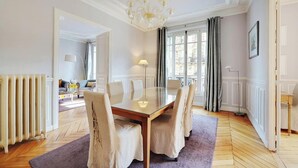 A dining area with a glass table, four chairs, a chandelier, and a view of the city through large windows. - NESTLED IN THE HISTORIC LATIN QUARTER,JUST STEPS FROM THE SEINE RIVER&NOTRE-DAME (Paris)
