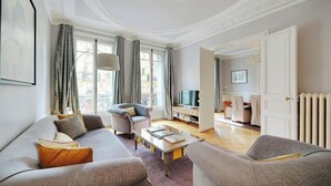 A living room with a sofa, armchair, and a coffee table. There is a television and a dining area visible through an open door. - NESTLED IN THE HISTORIC LATIN QUARTER,JUST STEPS FROM THE SEINE RIVER&NOTRE-DAME (Paris)