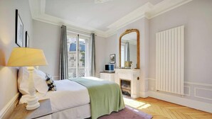 A bedroom with a bed, a bedside table with a lamp, a dresser, a fireplace, and a large mirror. - NESTLED IN THE HISTORIC LATIN QUARTER,JUST STEPS FROM THE SEINE RIVER&NOTRE-DAME (Paris)