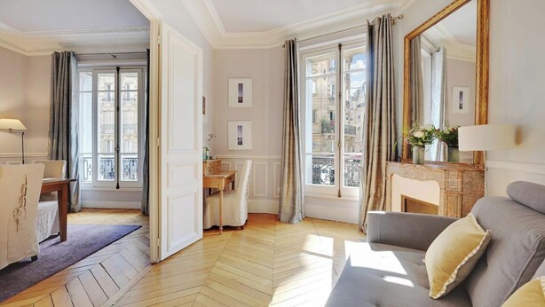 A well-lit room with a wooden parquet floor, a grey sofa, a wooden side table, a mirror, and a view of the city through large windows. - NESTLED IN THE HISTORIC LATIN QUARTER,JUST STEPS FROM THE SEINE RIVER&NOTRE-DAME (Paris)