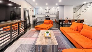 Una sala de estar moderna con un televisor grande, sofás naranja, una mesa de centro de madera y una cocina al fondo.