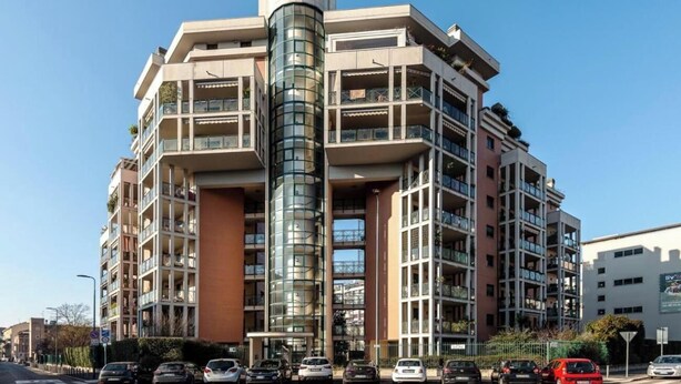 Un edifici residencial de diverses plantes amb balcons, una torre de vidre cilíndrica i un pàrquing ple de cotxes.