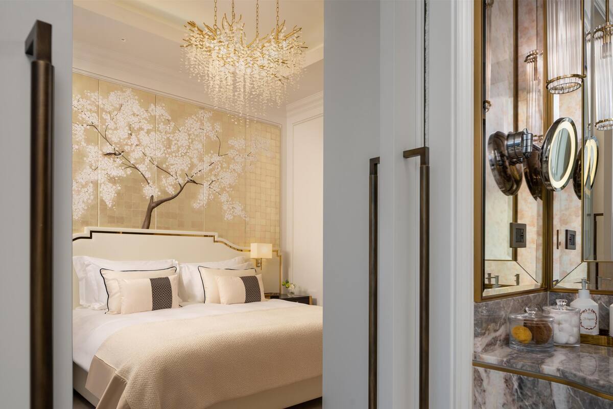 Une chambre d’hôtel avec un grand lit, un lustre, un mur décoré d’une fresque représentant un arbre, et une salle de bain avec un grand miroir et des articles de toilette.