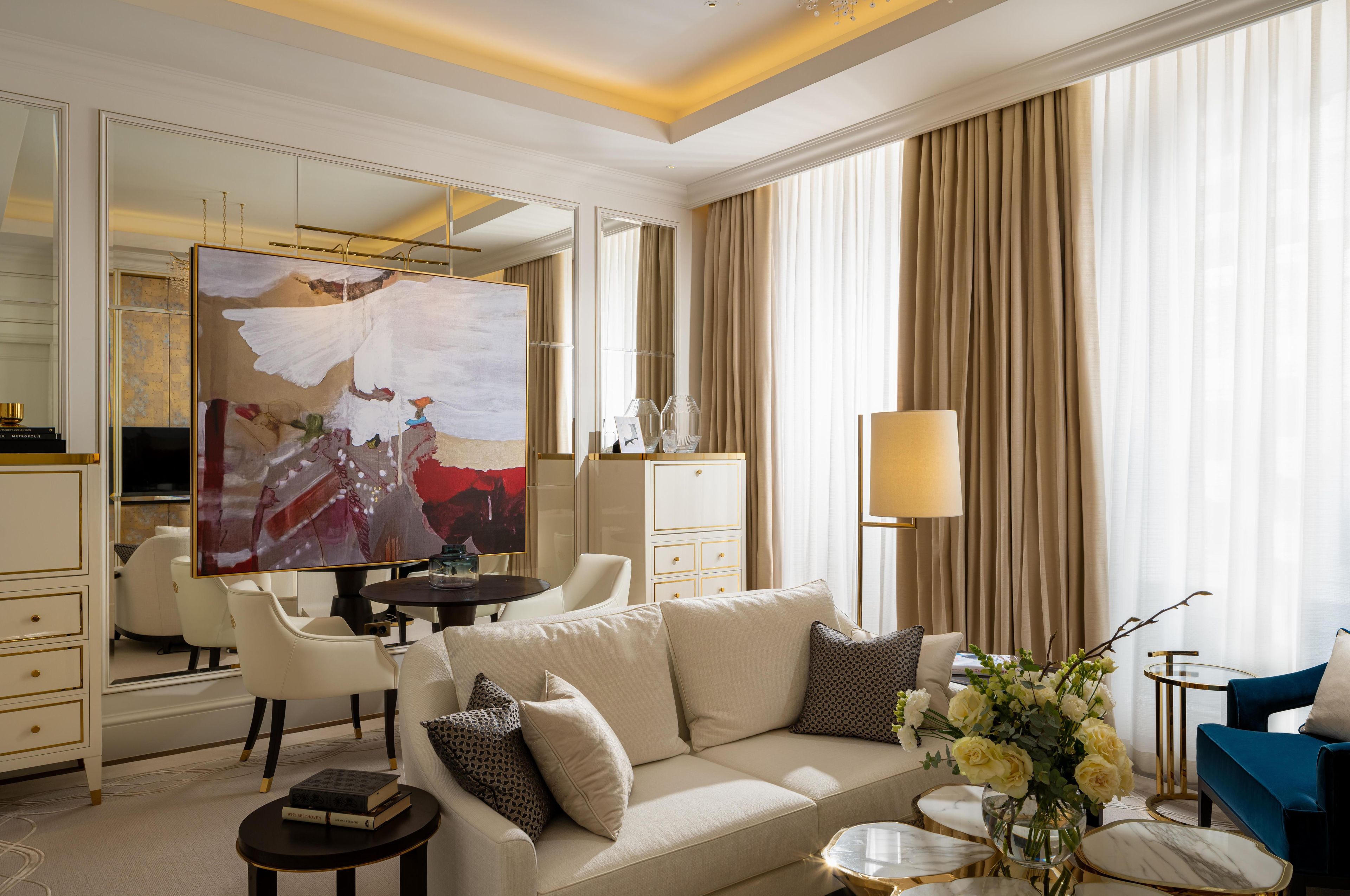 Una habitación de hotel moderna con un espejo grande, un sofá color beige, un sillón azul y un área de comedor con una mesa y sillas.
