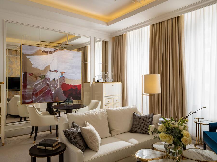 Una habitación de hotel moderna con un espejo grande, un sofá color beige, un sillón azul y un área de comedor con una mesa y sillas.