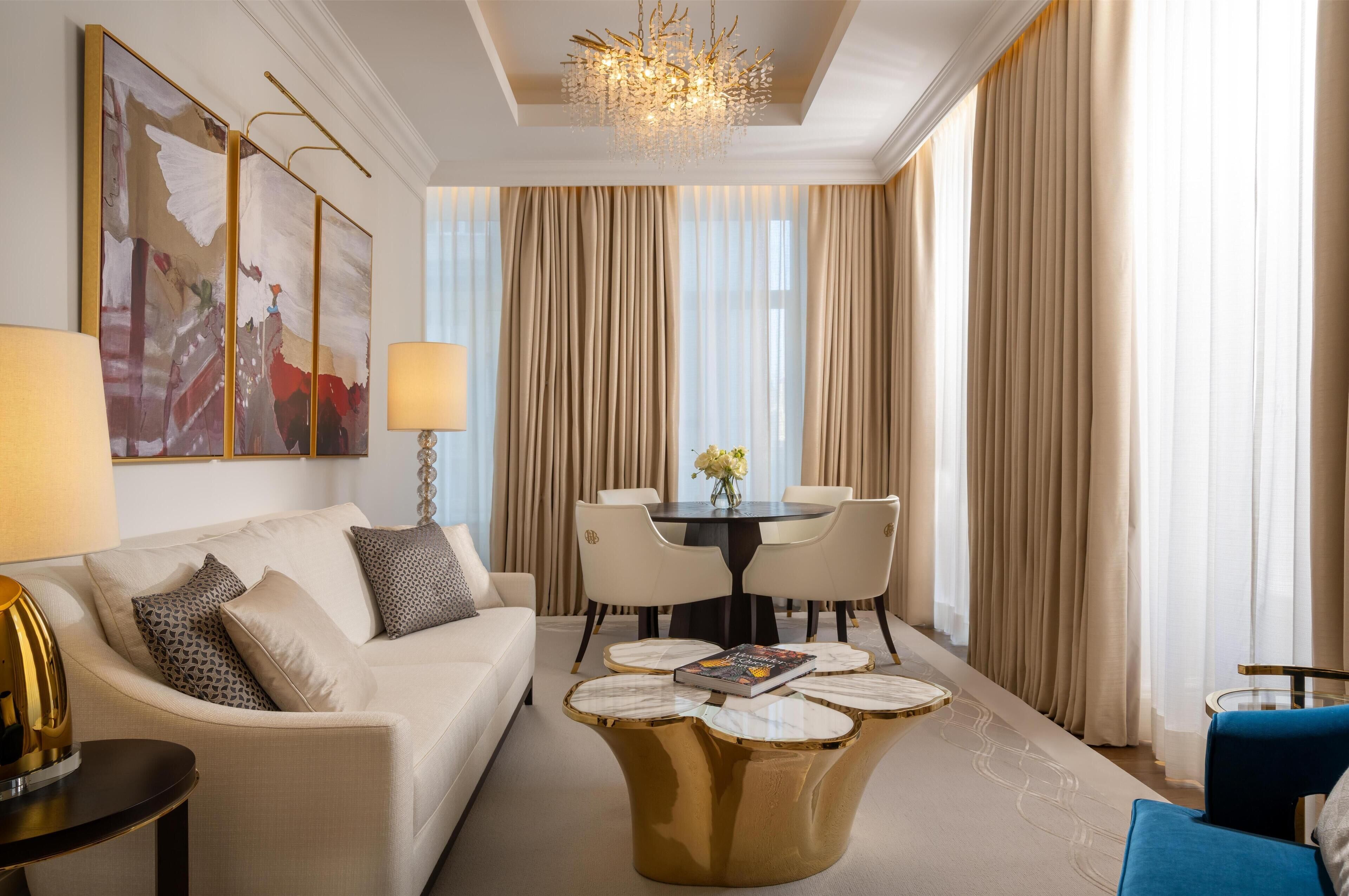 A modern hotel room with a white sofa, a round coffee table, a dining table with chairs, and a large window with curtains.