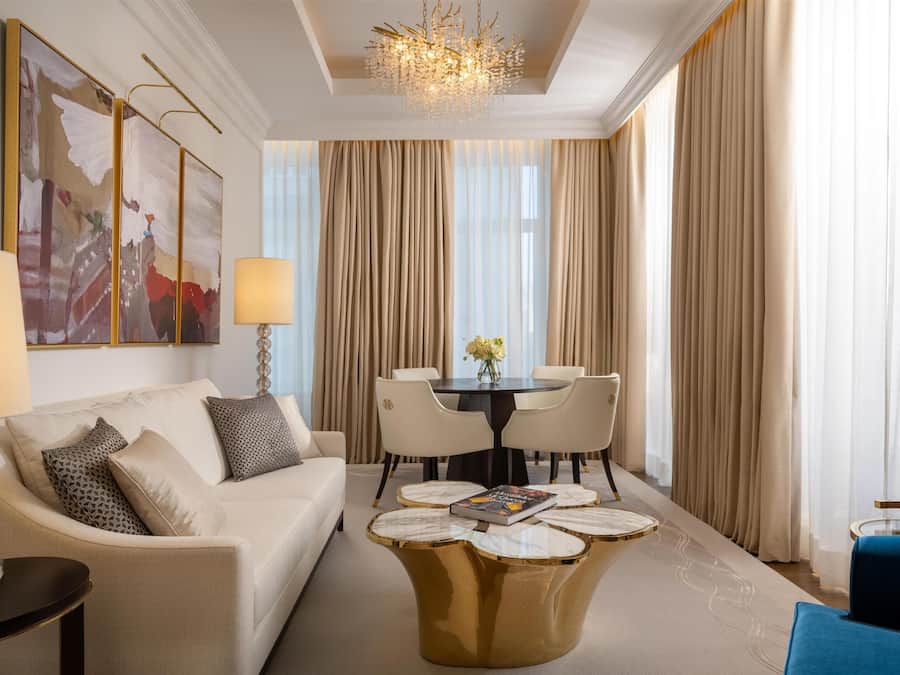 A modern hotel room with a white sofa, a round coffee table, a dining table with chairs, and a large window with curtains.