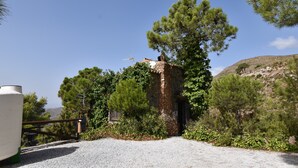 Un bâtiment rustique en pierre, doté d’une cheminée, entouré de pins et bordé d’un chemin de gravier.