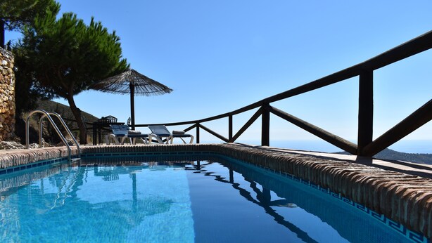 Une piscine d’un bleu éclatant, avec une terrasse en bois, des chaises longues et un mur en pierre.