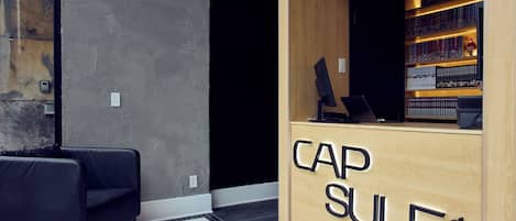 Un mostrador de recepción con detalles de madera y la inscripción "CAP SULE". A la izquierda, una silla de cuero negro.