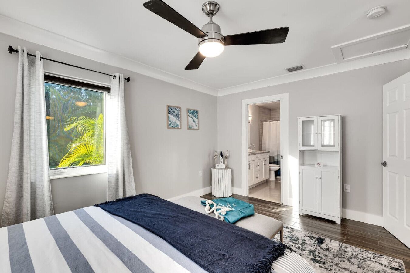 A bedroom with a bed, a ceiling fan, a window with curtains, and a view of a bathroom.