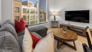 Een moderne woonkamer met een groot raam, een flatscreen televisie, een bank met rode en beige kussens, een kleine ronde tafel met een plant en een bijzettafeltje met een lamp.