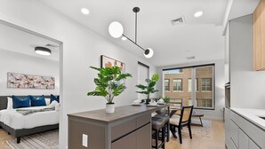 Een modern appartement met een centraal keukenblok, een eethoek en een slaapkamer met een groot raam.