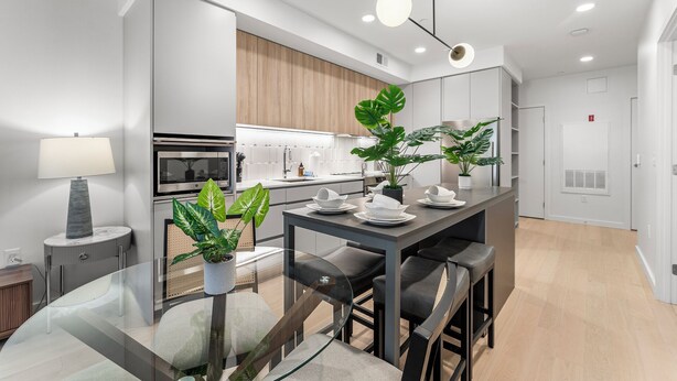 Een moderne keuken met een inbouwoven, magnetron en spoelbak. Er is een eethoek met een glazen tafel en stoelen, en potplanten.