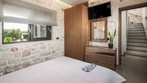 A bedroom with stone walls, a wooden wardrobe, a bed with a white bedspread, a mirror, and a TV mounted above the wardrobe.