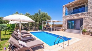 A swimming pool with lounge chairs and umbrellas, a stone building with a balcony, and a clear blue sky.
