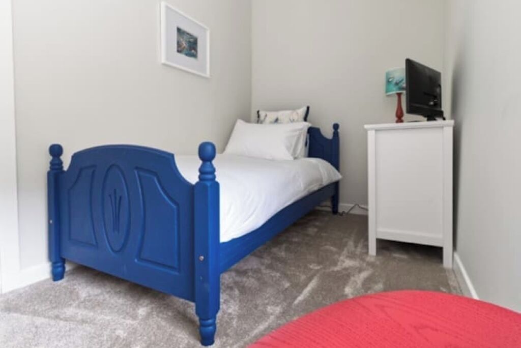 Un dormitorio con una cama azul, paredes blancas y un escritorio pequeño también blanco con un televisor.