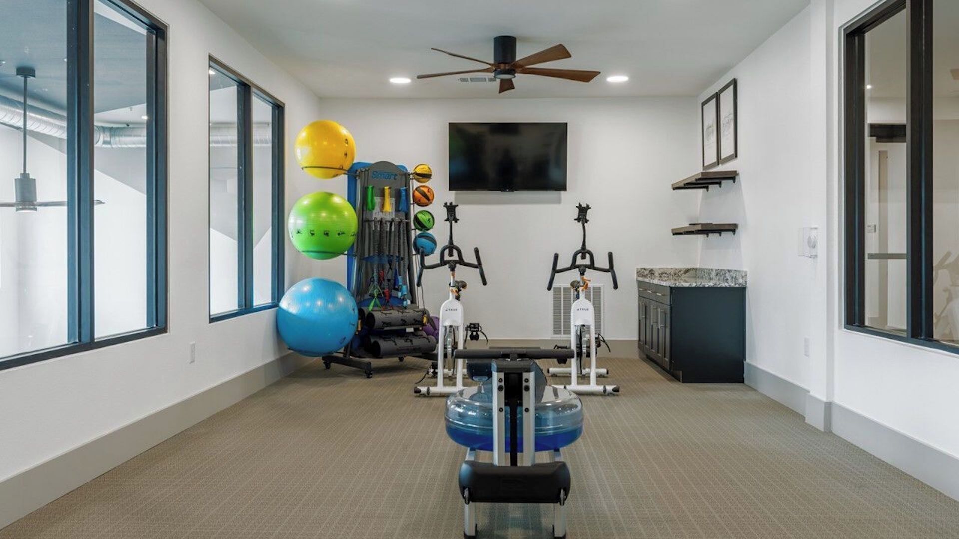 Ein Heimfitnessraum mit Trainingsgeräten, großen Fenstern und einem Deckenventilator.