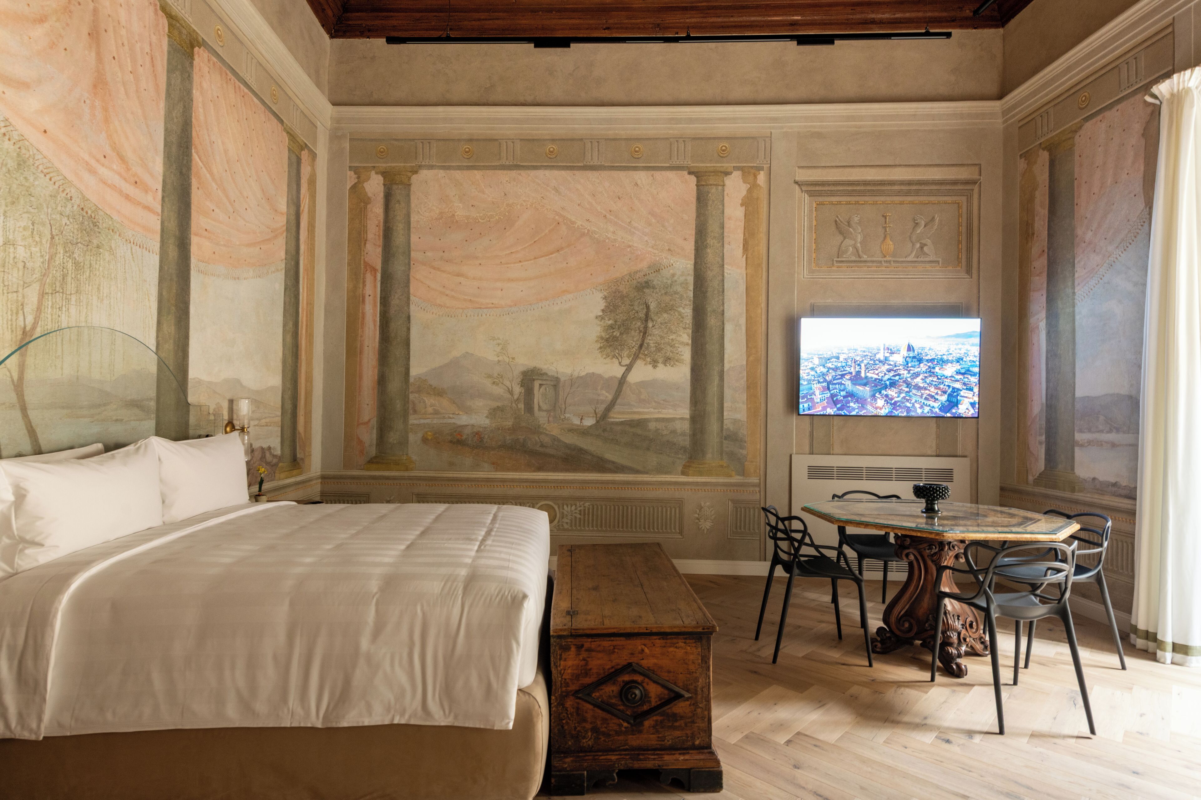 A bedroom with a bed, a wooden chest, a dining table with chairs, and a large mural on the wall.