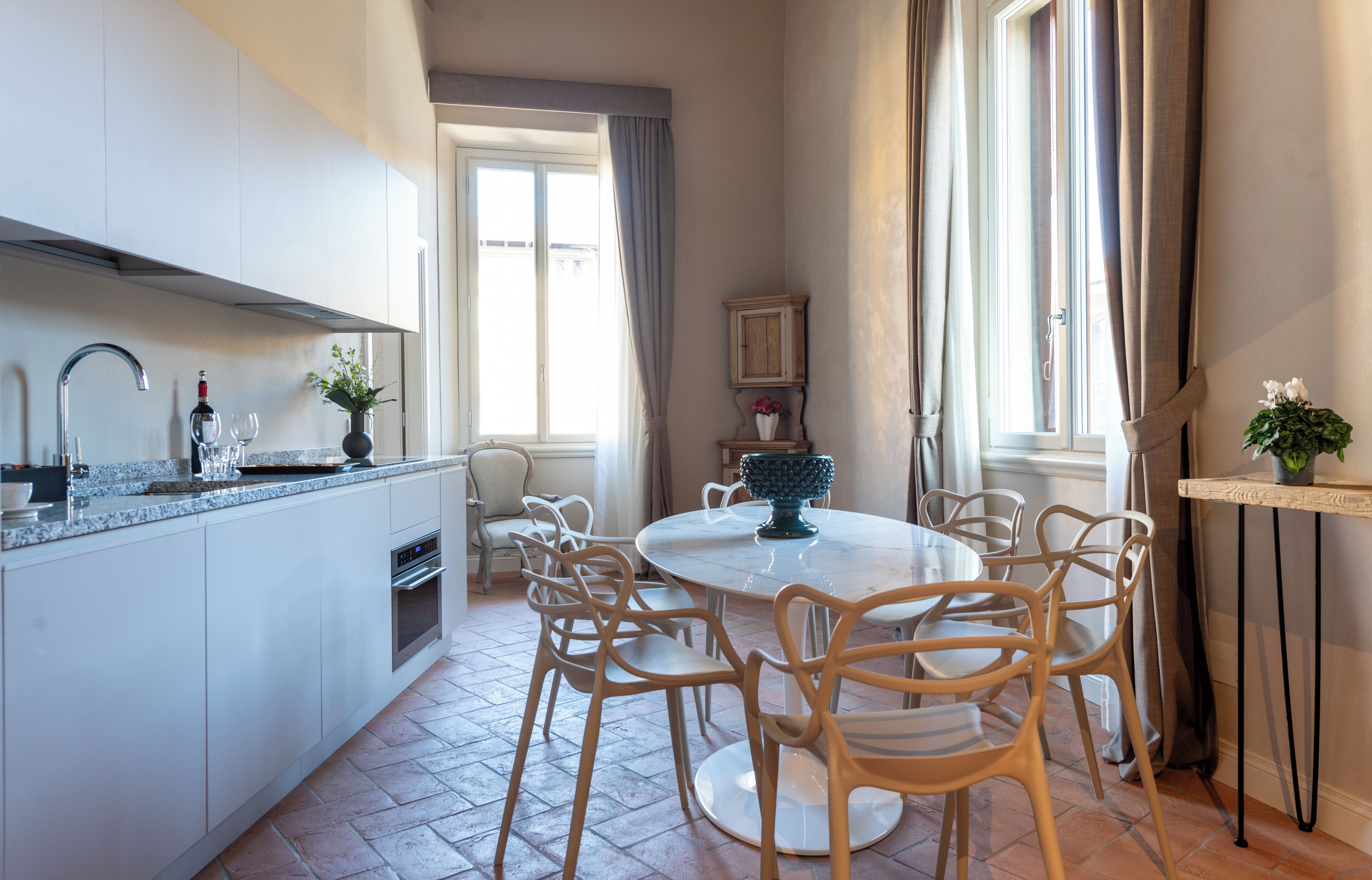 Eine moderne Küche mit weißen Schränken, einem runden Esstisch mit Stühlen, einem eingebauten Backofen und einem Fenster mit Vorhängen.