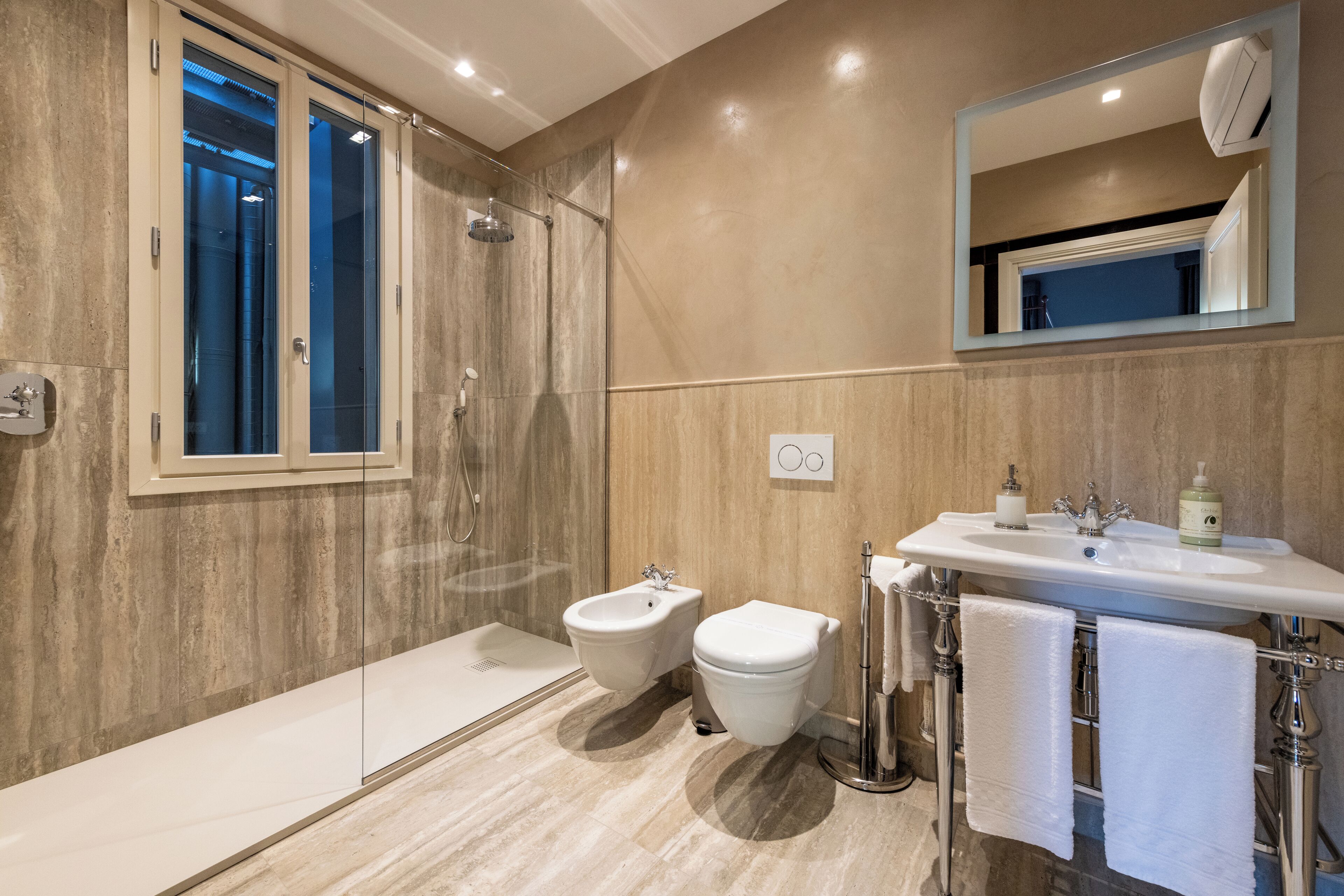 A modern bathroom with a glass shower enclosure, a bidet, a sink with a mirror, and two towels hanging on the wall.
