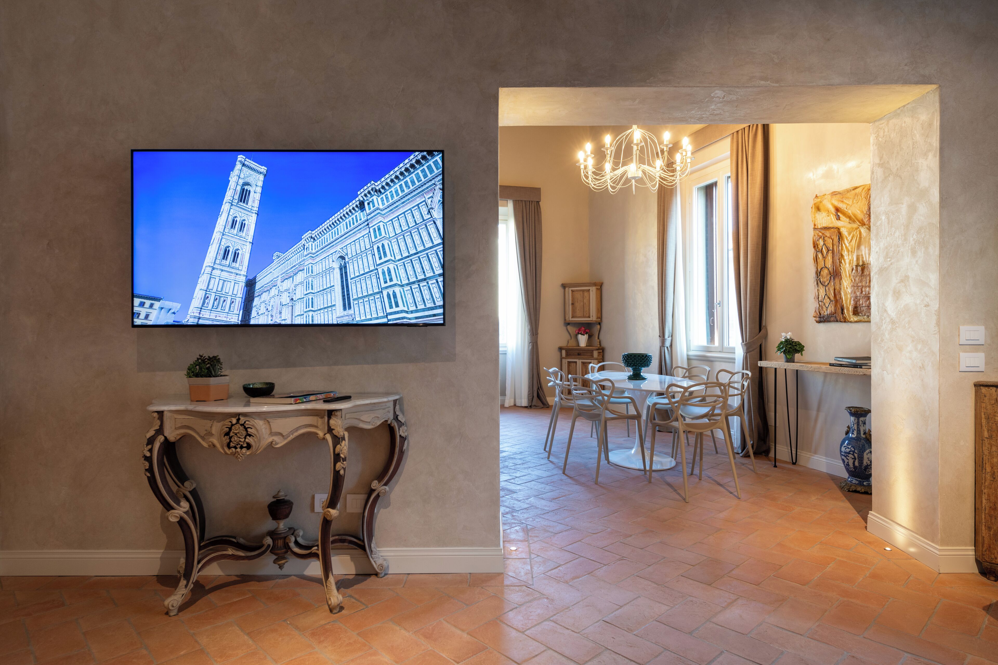 Een kamer met een flatscreen televisie waarop een gebouw te zien is, een eethoek met een kroonluchter en een tafel met stoelen.