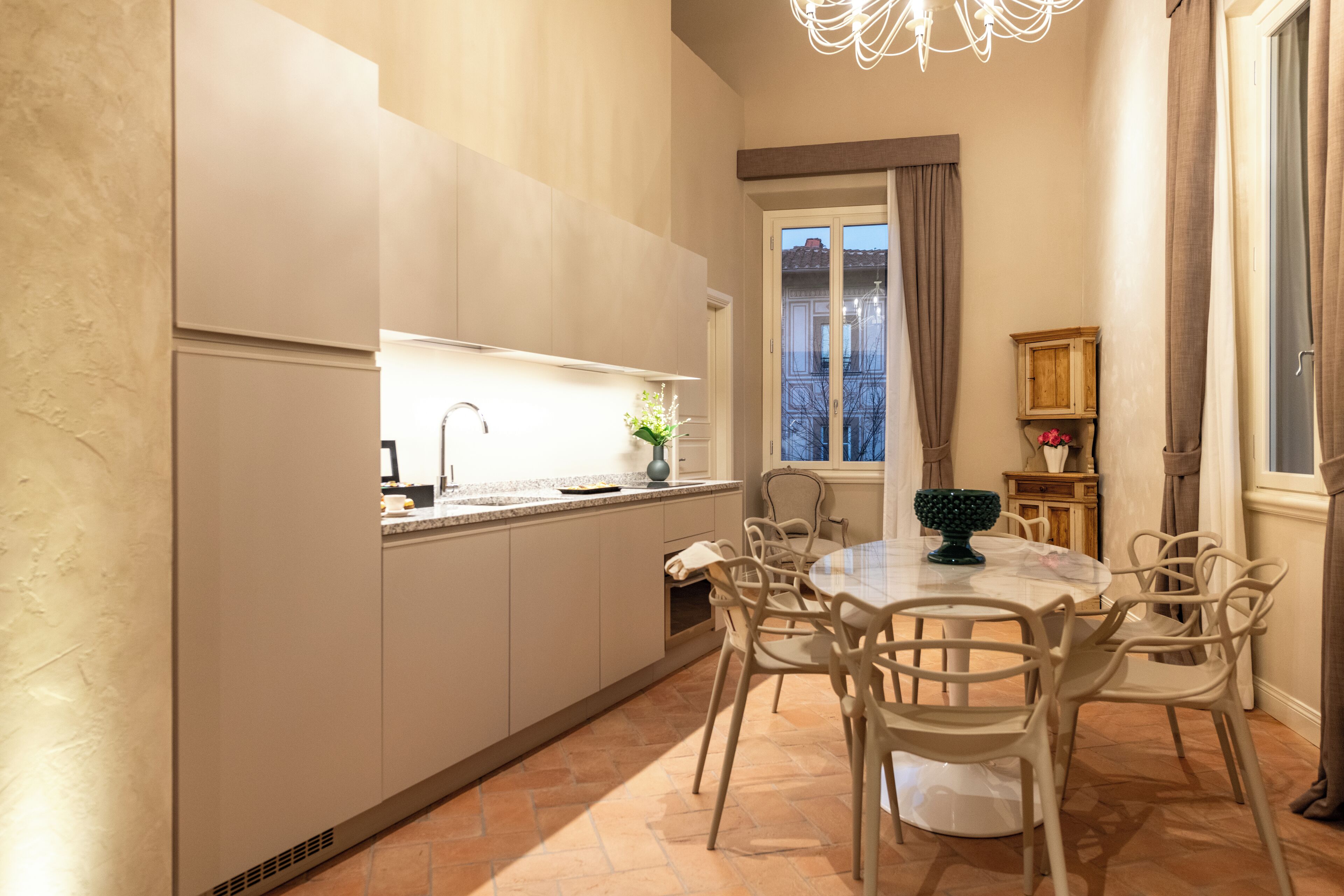 A modern kitchen with a dining area, featuring a round table and chairs, a built-in oven, and a window with curtains.