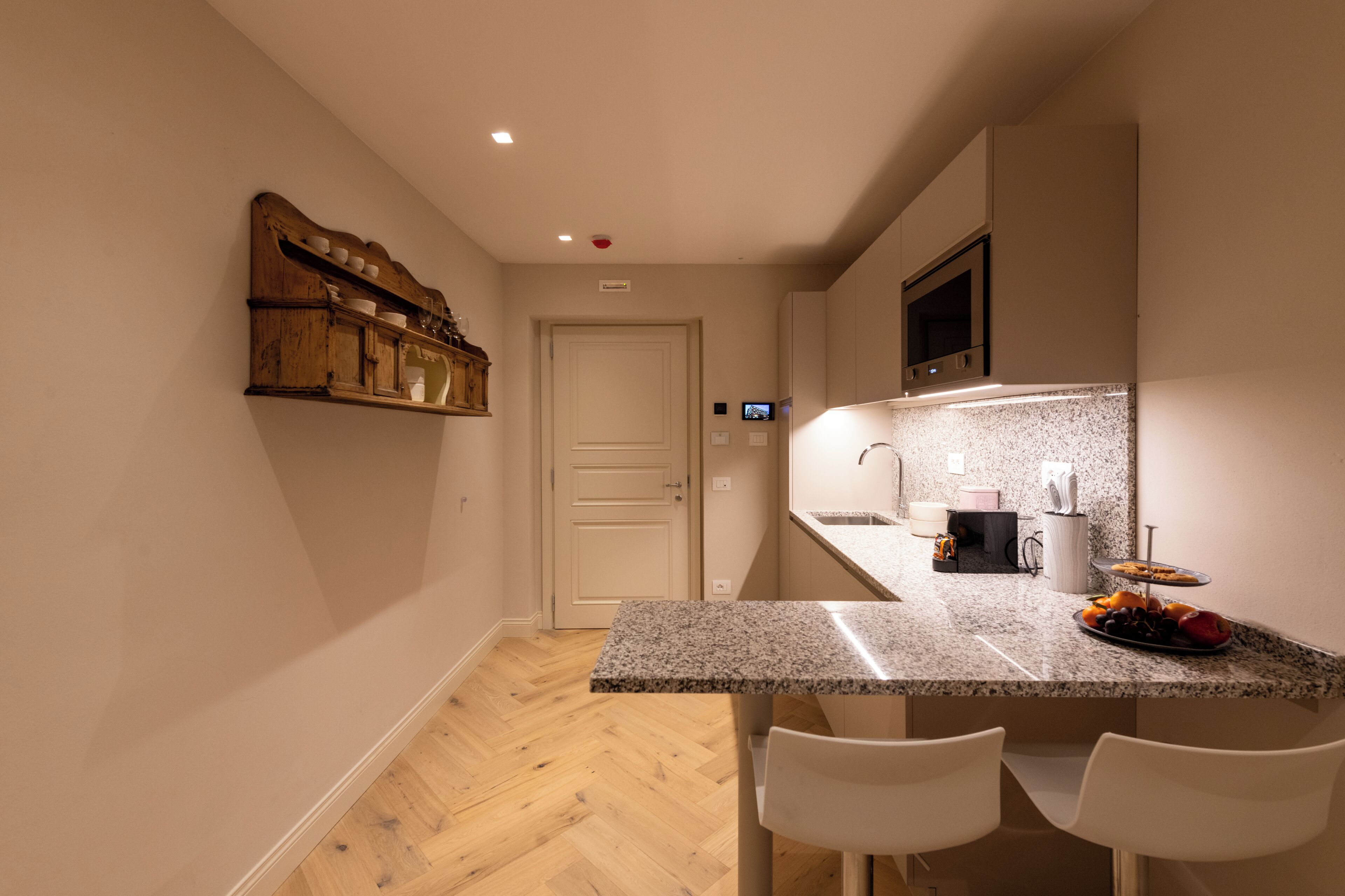 A compact kitchen with a granite countertop, two white chairs, a wooden shelf with dishes, and a microwave above the counter.