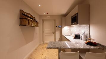 A compact kitchen with a granite countertop, two white chairs, a wooden shelf with dishes, and a microwave above the counter.