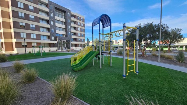 A playground with a slide and climbing structures, surrounded by artificial grass and a building in the background.