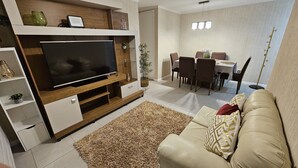 A modern living room with a flat-screen TV, a beige sofa, a dining area with a table and chairs, and a small plant.