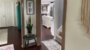 Un pasillo con una planta en maceta, un cuadro enmarcado y una alfombra que conduce a una zona de cocina con armarios blancos y un refrigerador.