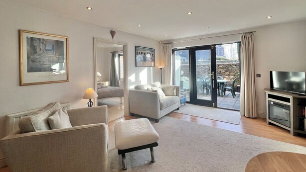 A modern living room with a sofa, armchair, and ottoman, a television, and a sliding glass door leading to an outdoor area with chairs. - Central Spacious Ground Floor Flat with Courtyard (Kingsbridge)