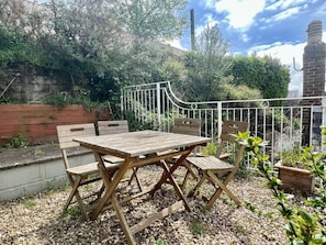 A wooden table and chairs set outdoors with a metal fence and greenery in the background.