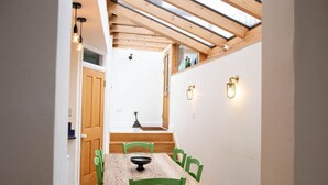 A narrow hallway leading to a dining area with a wooden table and green chairs, a wooden staircase, and a skylight above.