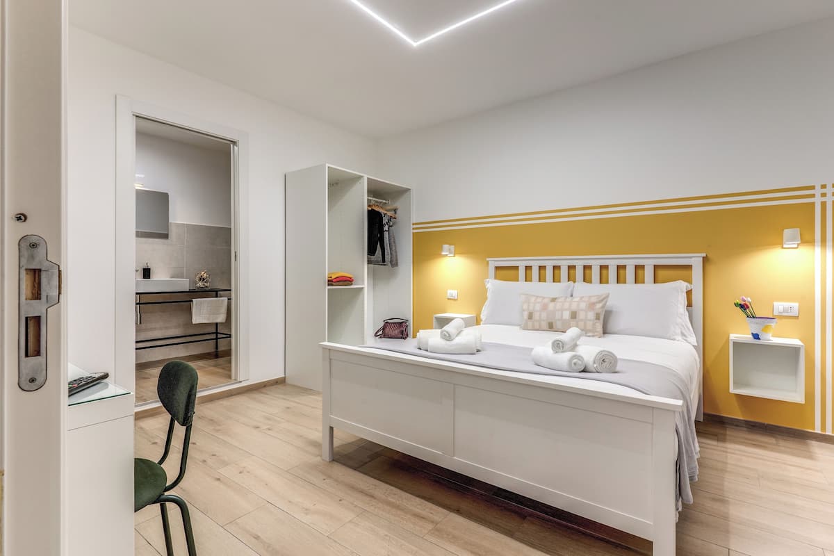 A modern hotel room with a bed, bedside tables, a wardrobe, and a bathroom visible through an open door.