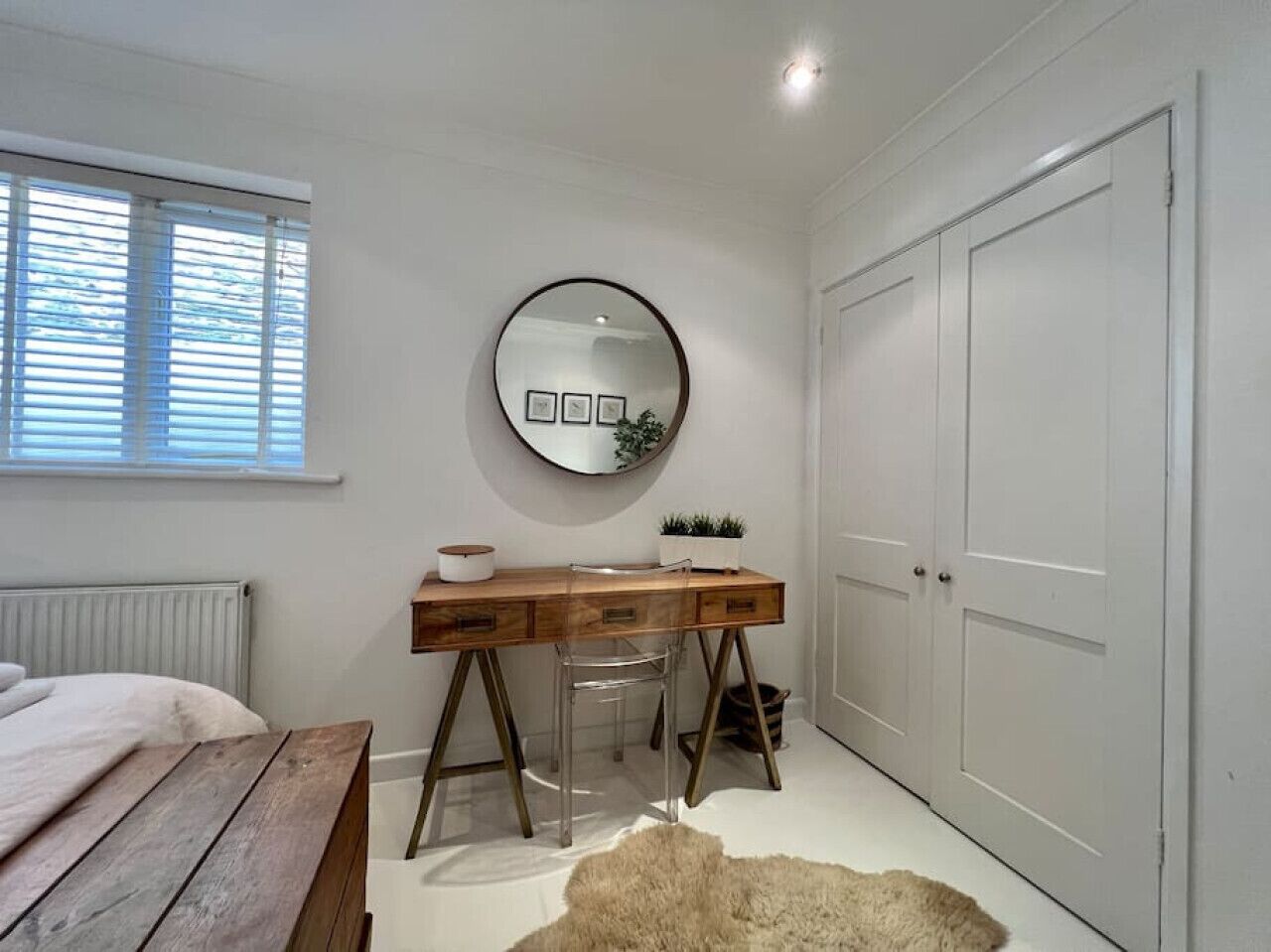 A bedroom with a wooden desk, a round mirror, a white radiator, a window with blinds, and a white door.