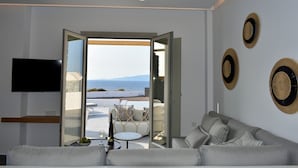 A modern living room with a grey sofa, a flat-screen TV mounted on the wall, and a glass door leading to an outdoor seating area with a view of the sea.