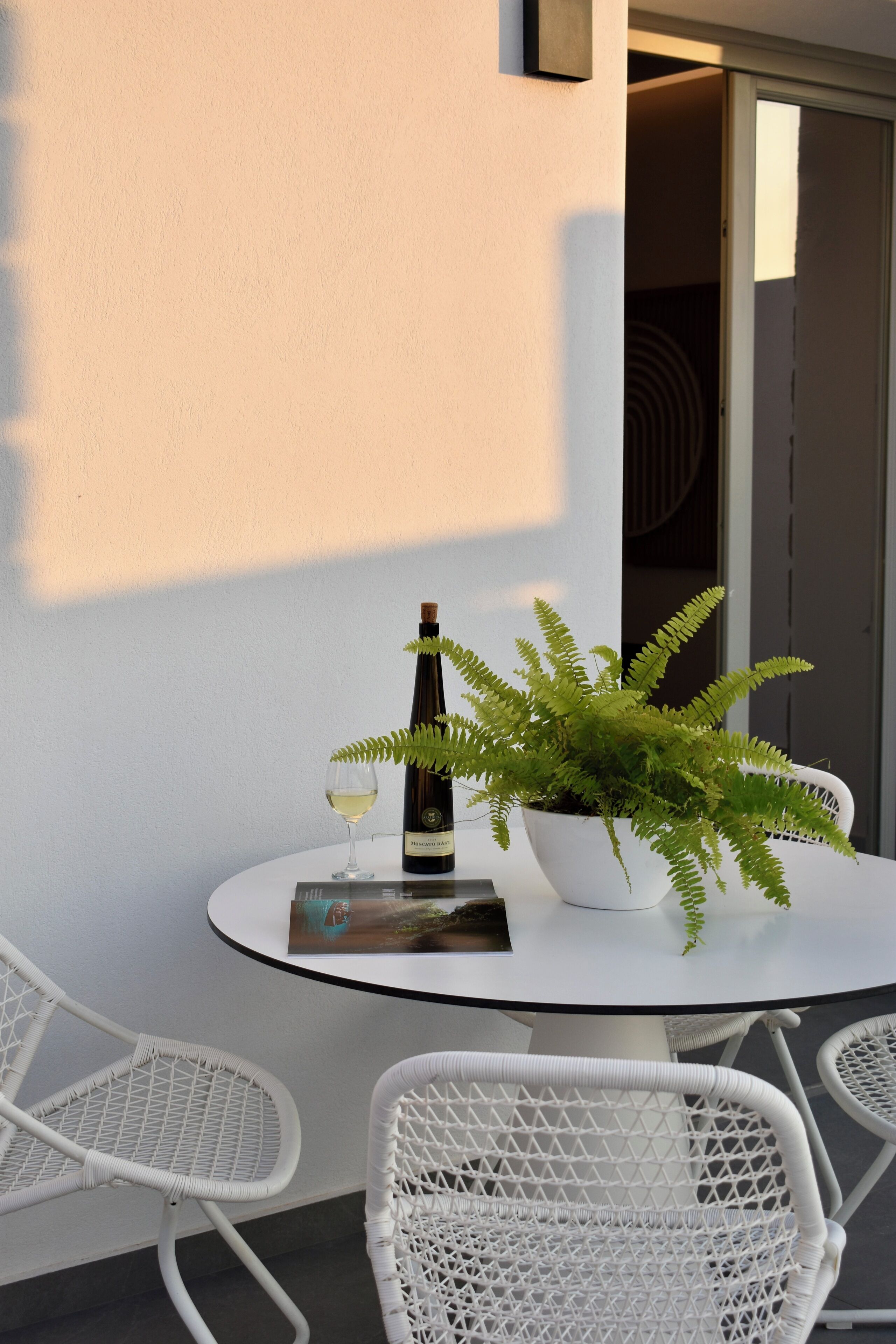 A round table with a bottle of wine, a glass, and a potted fern. There are two white wicker chairs around the table.