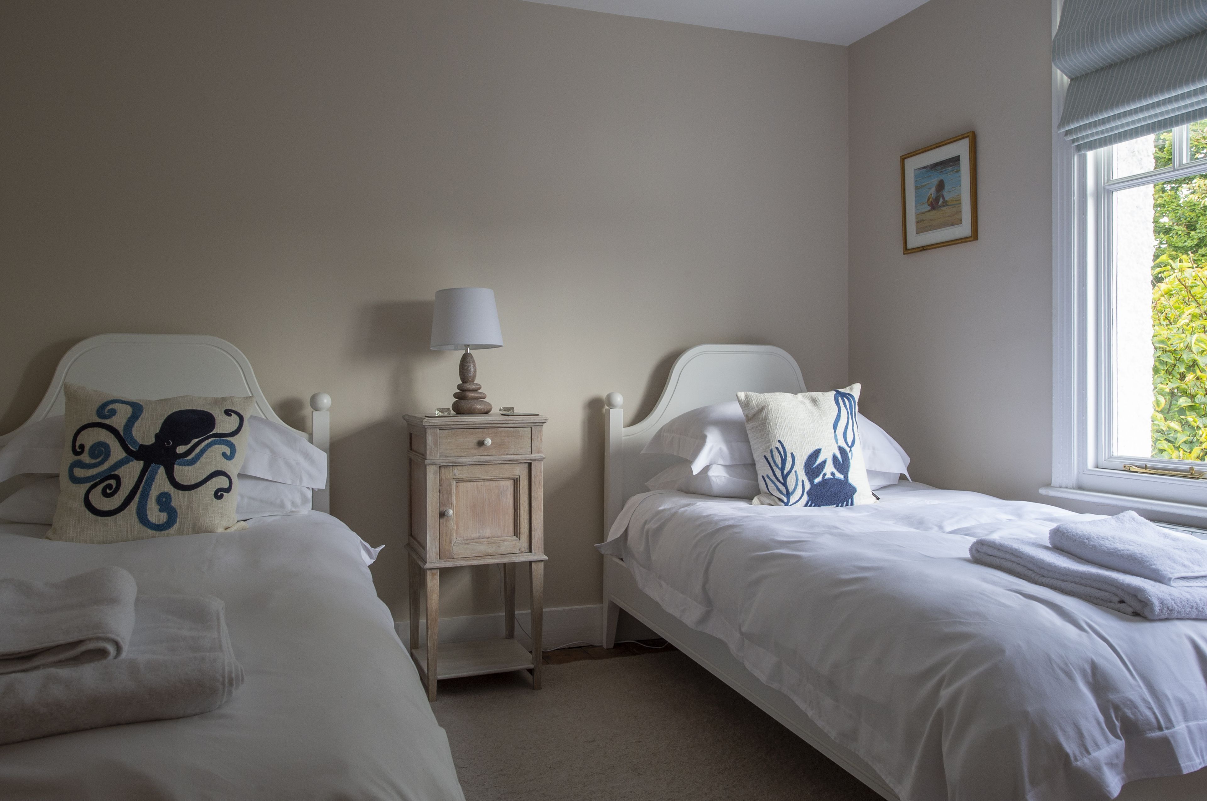 A bedroom with two beds, a nightstand with a lamp, and a window with a view of greenery.