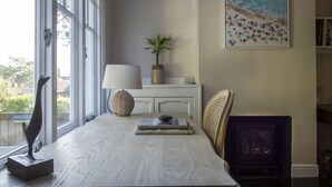 Een kamer met een groot raam, een open haard, een tafel met boeken, een lamp en een plant.