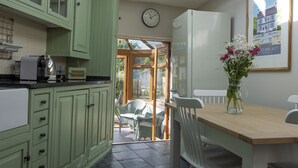Een keuken met groene kastjes, een witte koelkast en een eettafel met stoelen. Er hangt een klok aan de muur en een lijst met een foto boven de tafel.