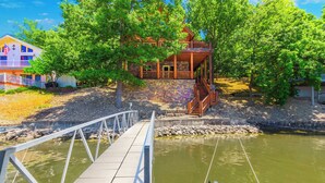 A wooden cabin with a stone foundation, a balcony, and a dock extending over a body of water.