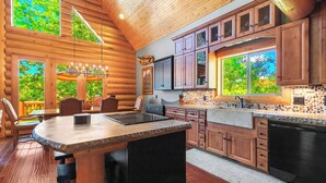 A log cabin interior with a wooden dining area, kitchen with stone countertop, and large windows offering views of greenery.
