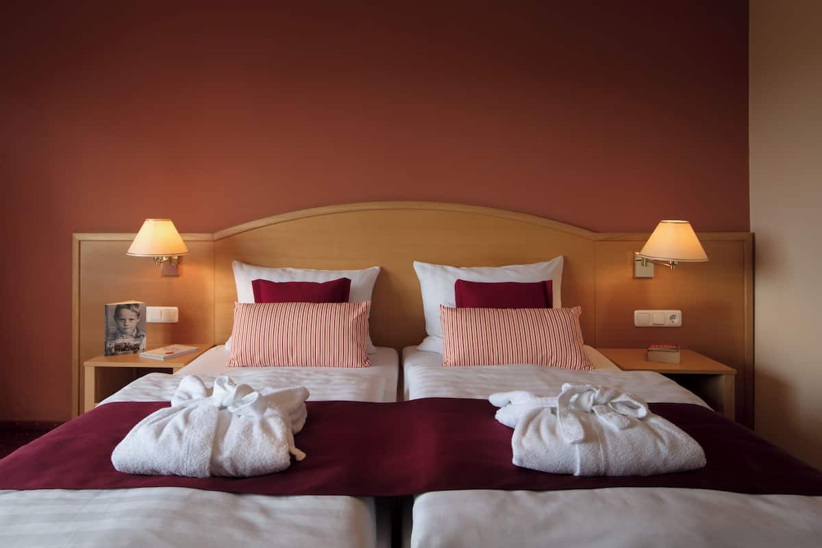 Habitación de hotel con dos camas, cada una con ropa de cama blanca y almohadas rojas. Dos lámparas de noche, una mesita de noche con lámpara, y una manta roja en cada cama.