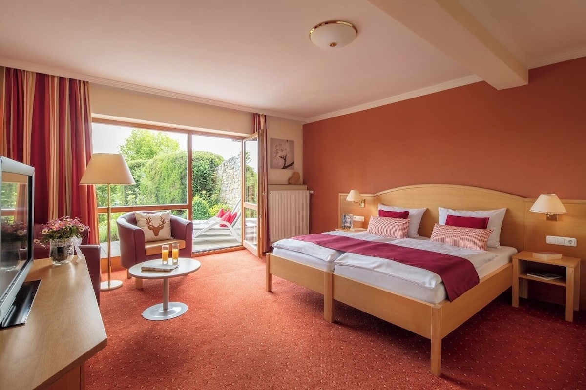 Habitación de hotel con cama, televisor, escritorio, silla, una pequeña mesa con bebidas y balcón con vistas a zonas verdes.