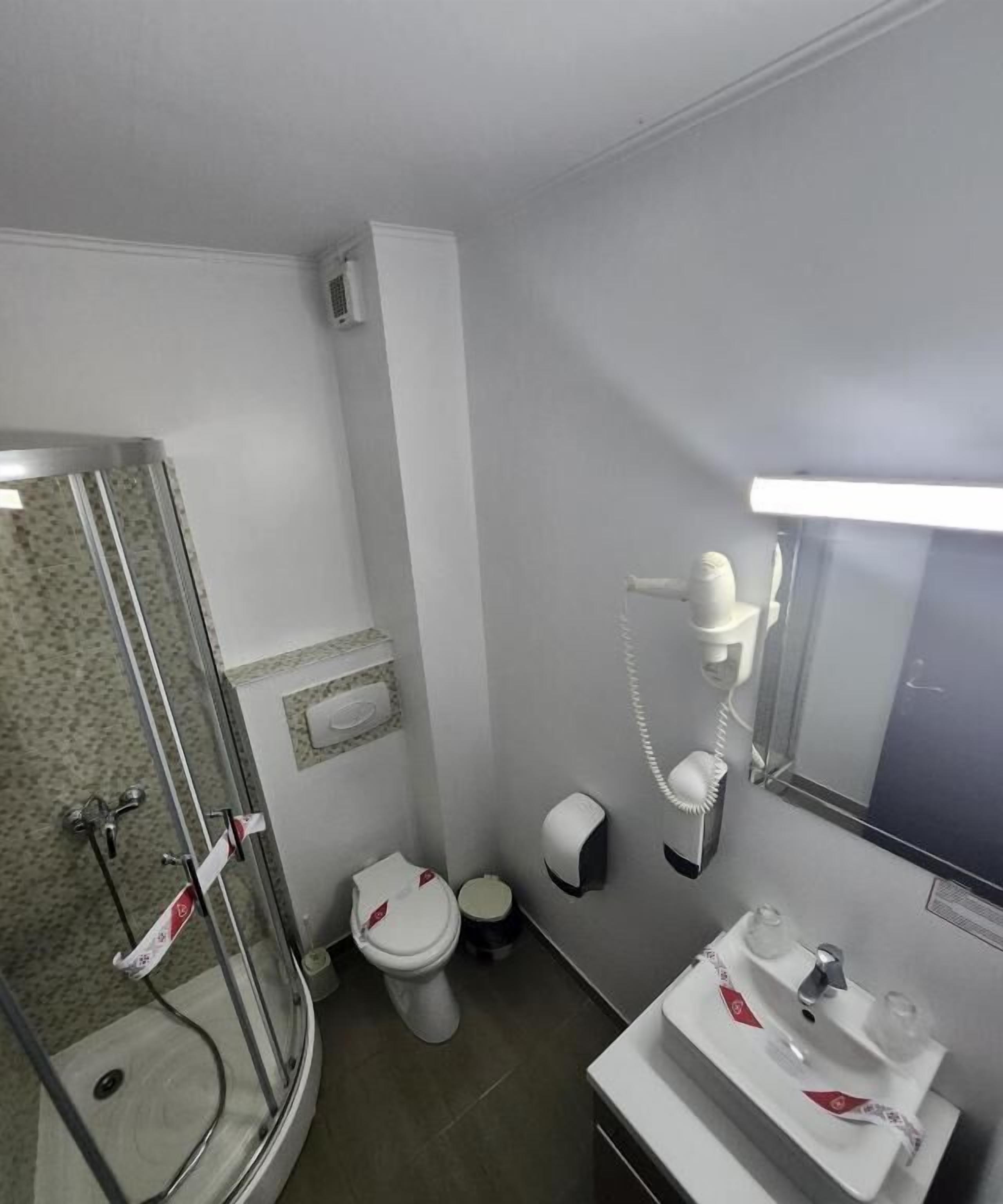 Uma casa de banho compacta com cabine de duche em vidro, sanita, lavatório com espelho e secador de mãos.