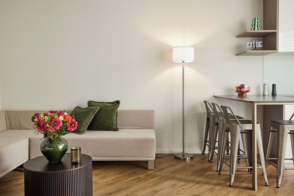 A modern living room with a sofa, a round table with a vase of flowers, a floor lamp, and a dining area with bar stools.